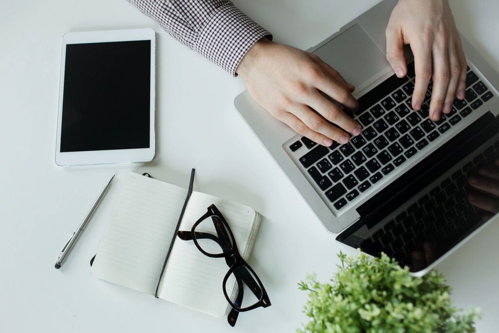 A person's hands typing on a laptop keyboard with a tablet and notepad nearby on a desk.