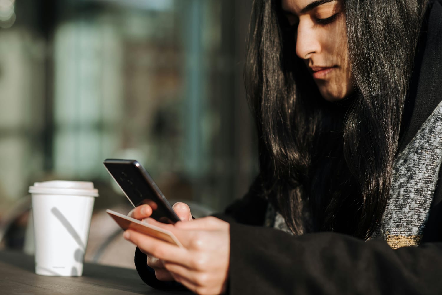 A woman with long dark hair using a smartphone while holding a card and a coffee cup on a table.