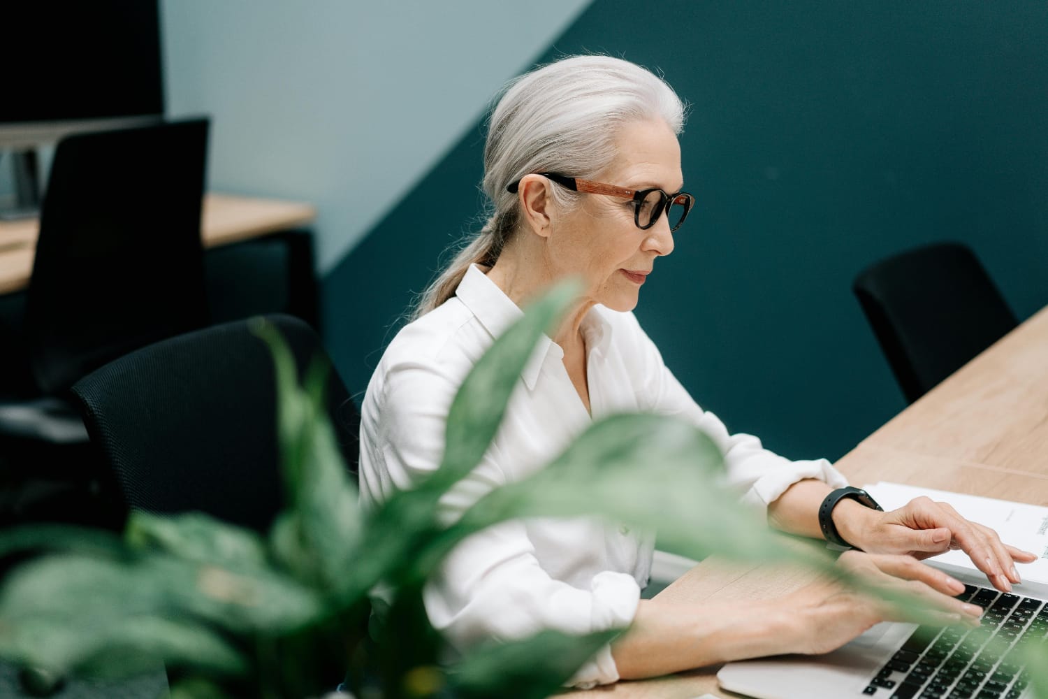 A woman with grey hair and glasses typing on a laptop in an office setting.