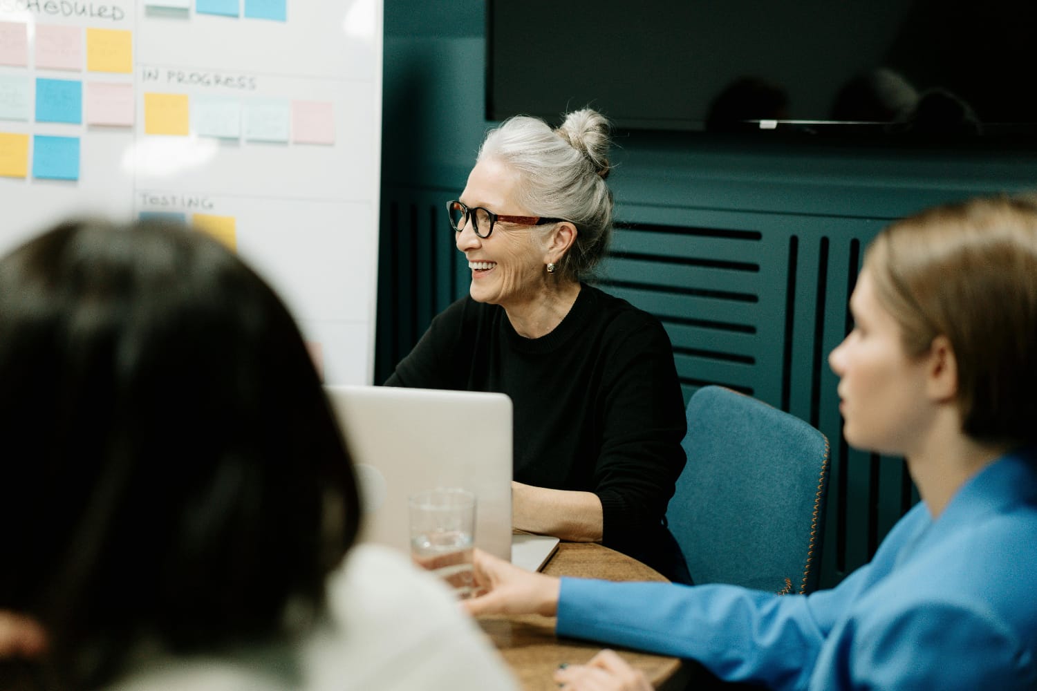 A woman with grey hair and glasses smiling while seated at a table with a laptop during a meeting.
