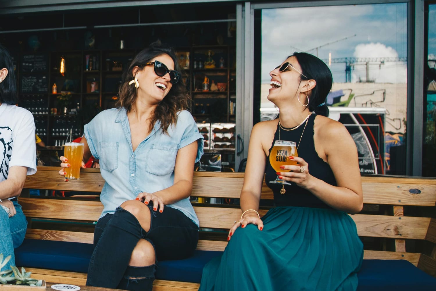 Two women sitting on a bench, laughing and holding drinks in a casual setting.