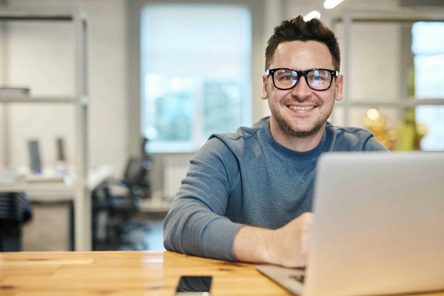 Man wearing glasses smiling while using a laptop in an office setting with wooden desk.