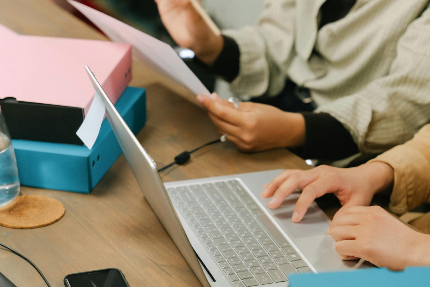 Two hands typing on a silver laptop with documents and folders on a wooden table.