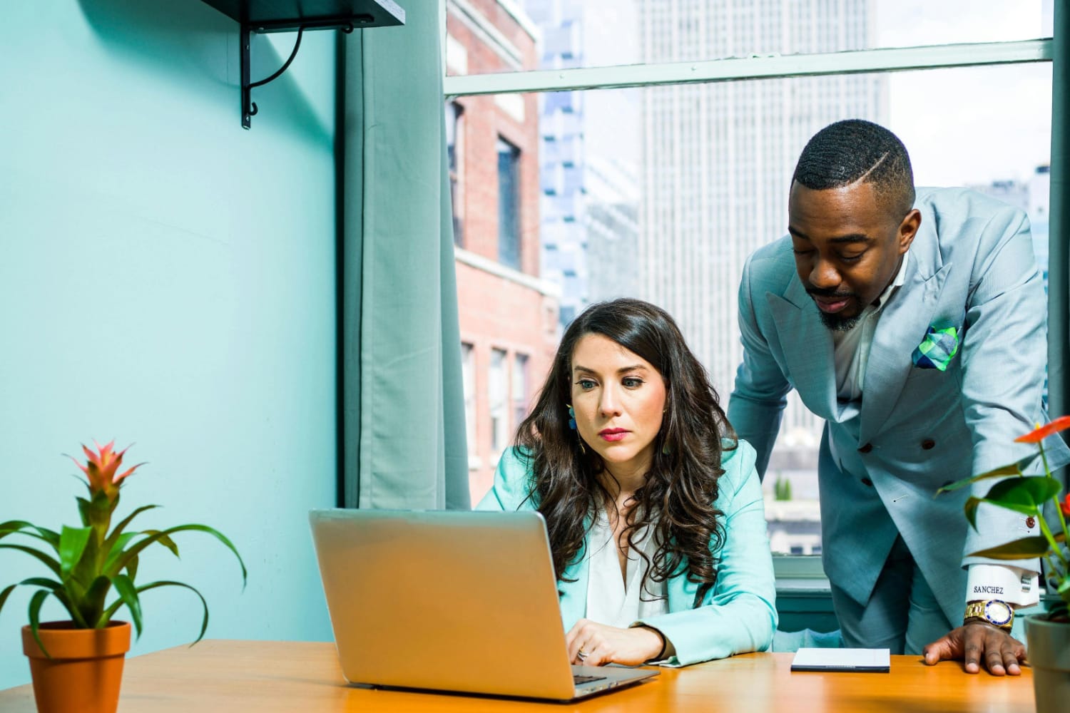 A woman working on a laptop while a man stands beside her in a professional setting.