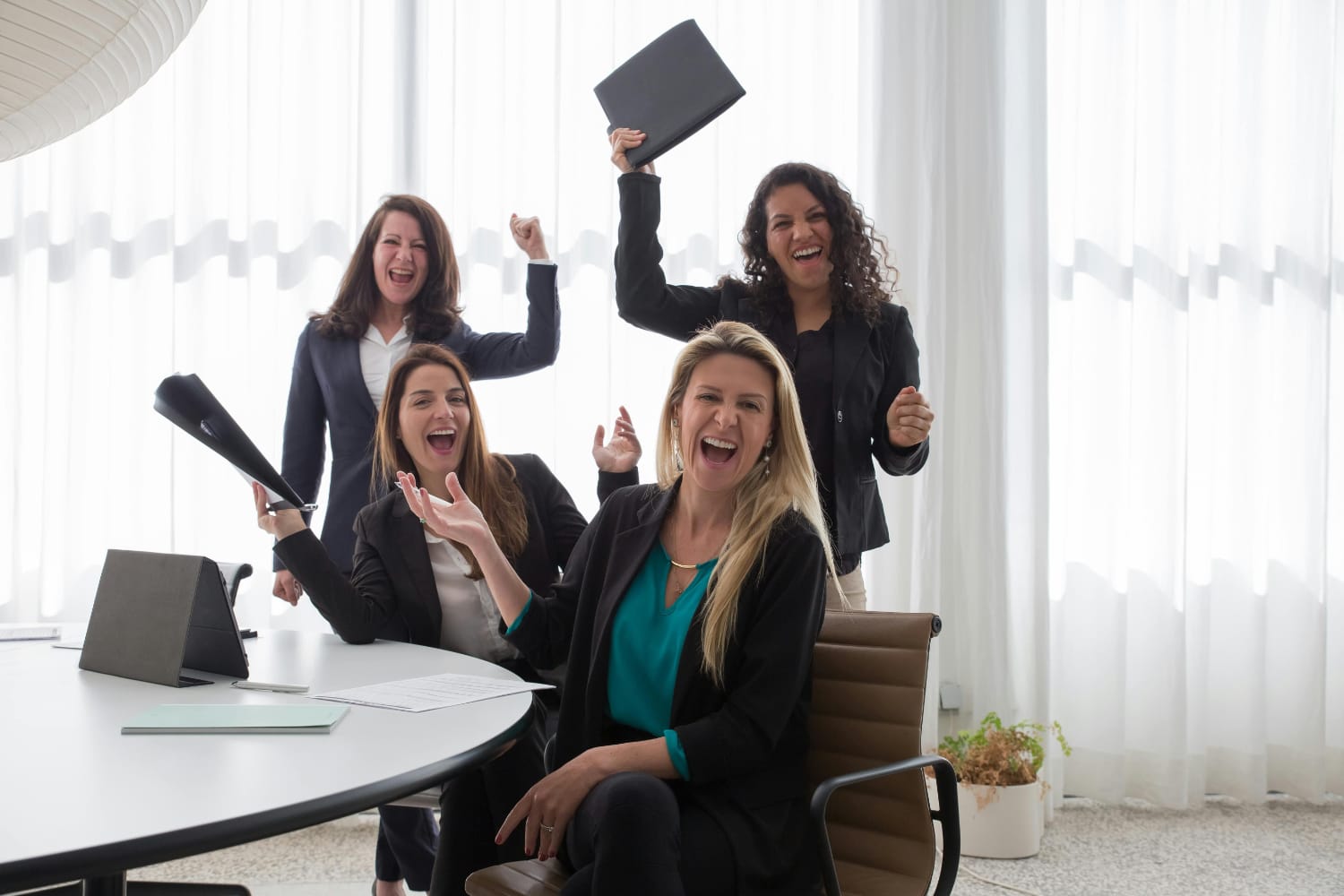 Four women in business attire celebrating with documents and tablets in an office environment.