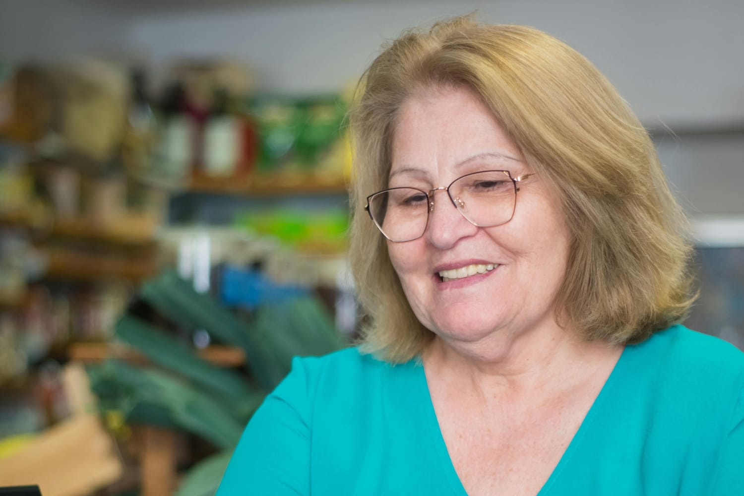 Older woman with glasses and blonde hair smiling while looking down, wearing a turquoise top.