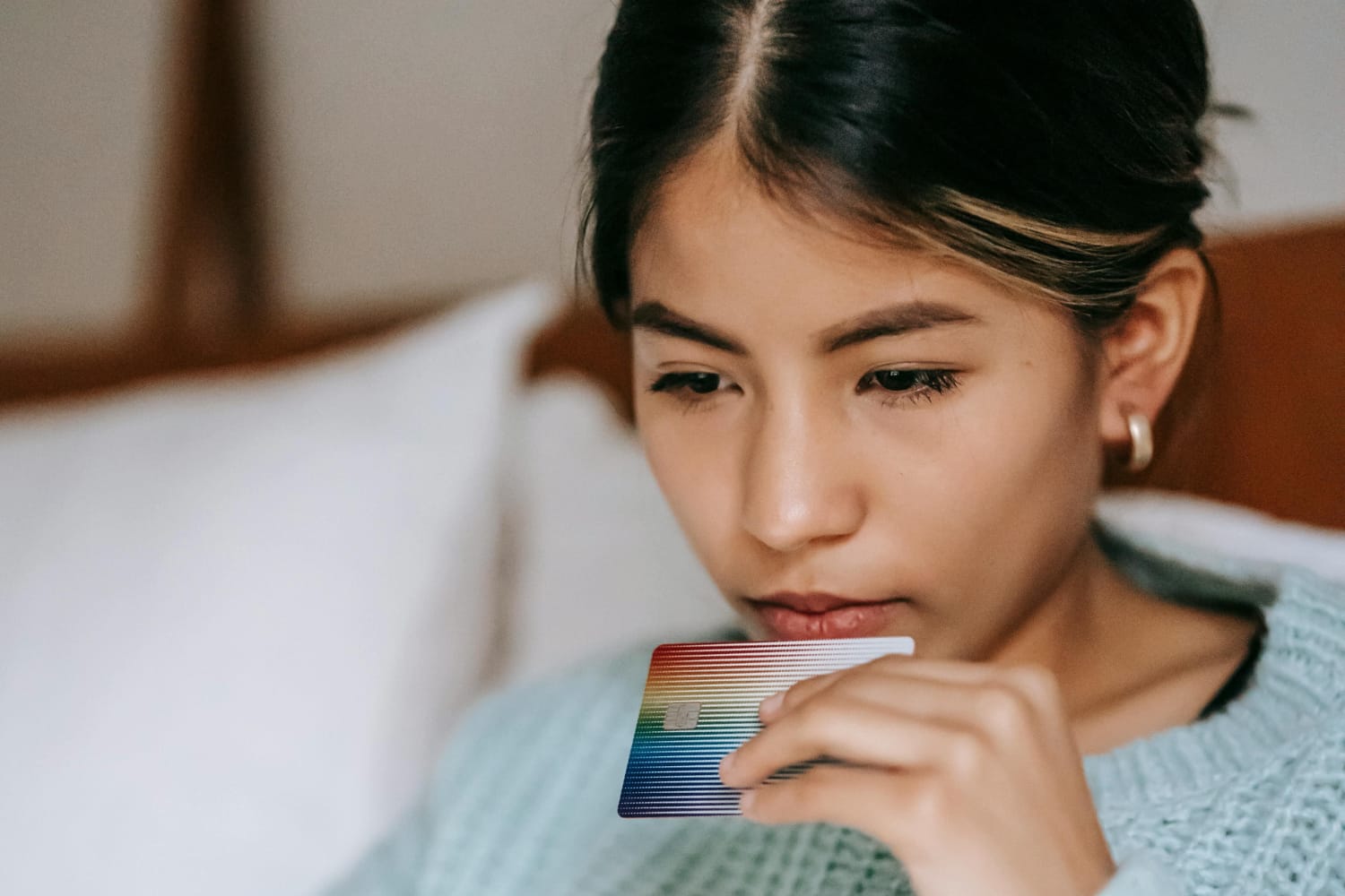 A woman with long hair holding a credit card near her mouth while looking down thoughtfully.