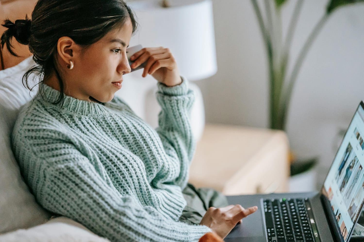 Woman with long hair in a green sweater using a laptop while sitting on a sofa.