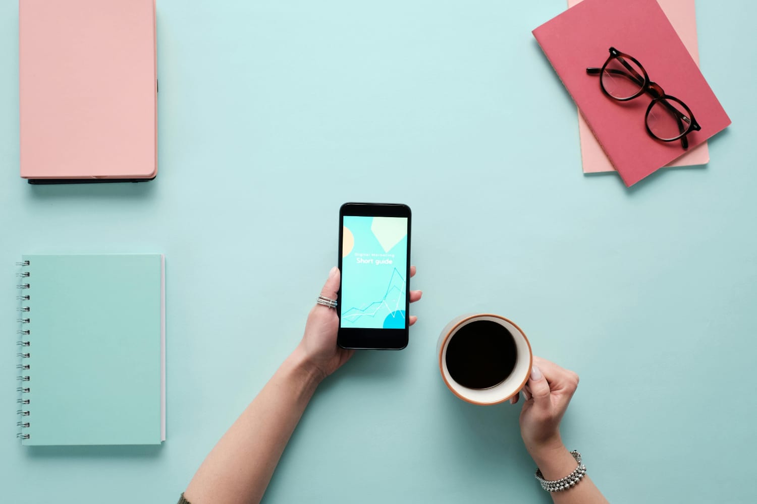 A hand holding a smartphone displaying a guide, with a cup of coffee and stationery on a light blue surface.