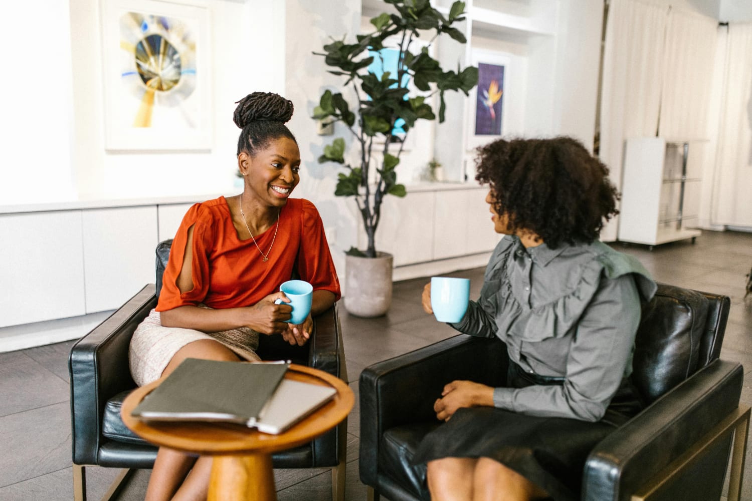 Two women sitting in armchairs, holding coffee cups and engaging in conversation indoors.