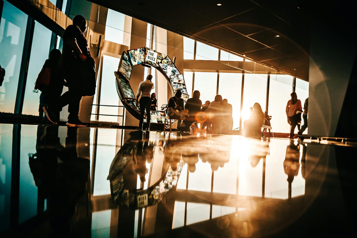 Silhouetted figures walking and sitting in a brightly lit modern indoor area with reflective flooring.