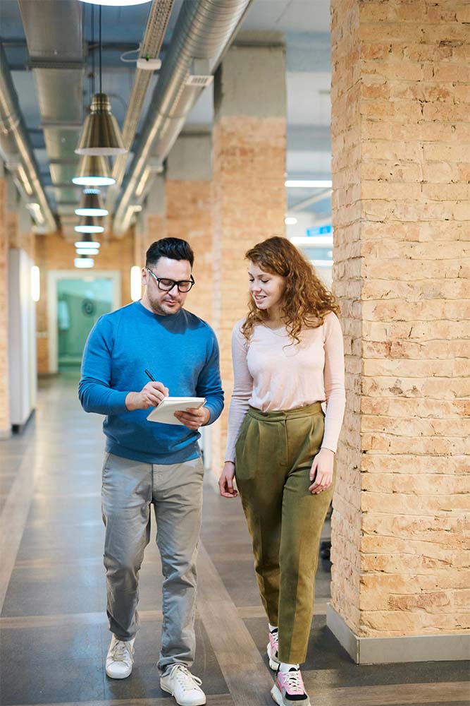 A man and a woman walking together in an office corridor, the man holding a notepad.