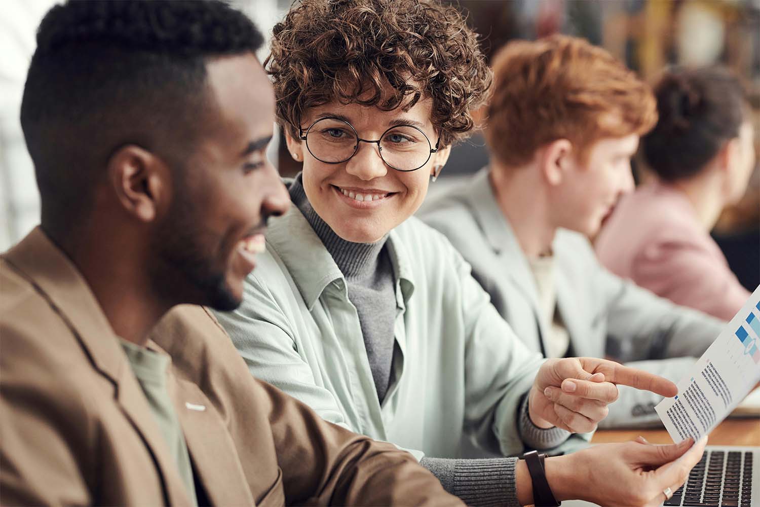 Two individuals engaged in conversation while others work at a table in a collaborative environment.