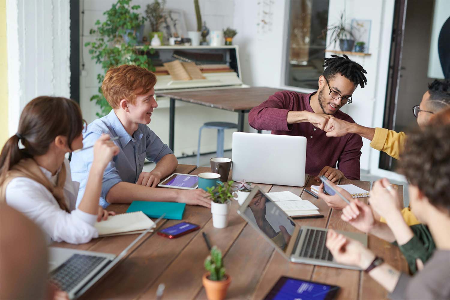 A diverse group of individuals participating in a meeting around a wooden table with laptops and notebooks.