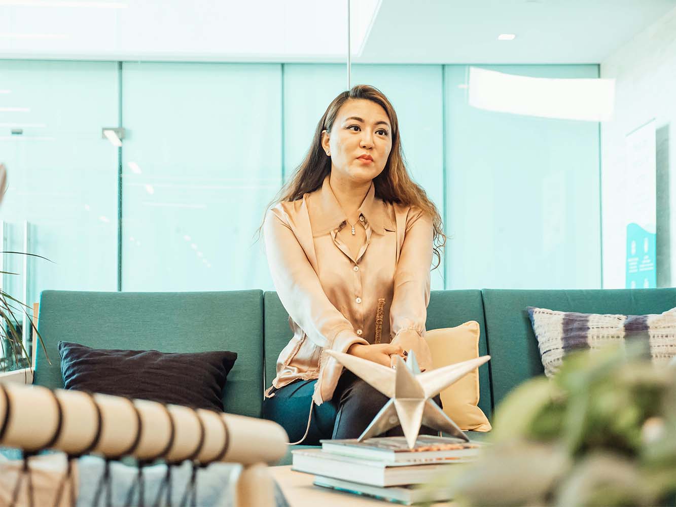 A woman in a light blouse sitting on a teal sofa with decorative cushions in an office environment.