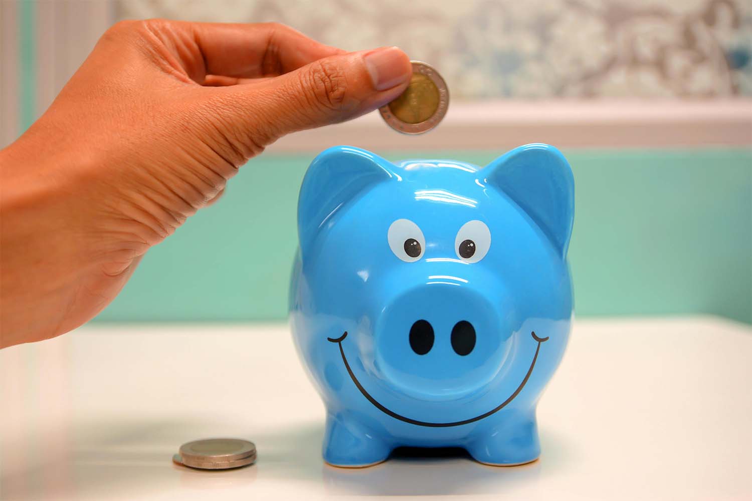 Hand placing a coin into a blue piggy bank with a smiling face.