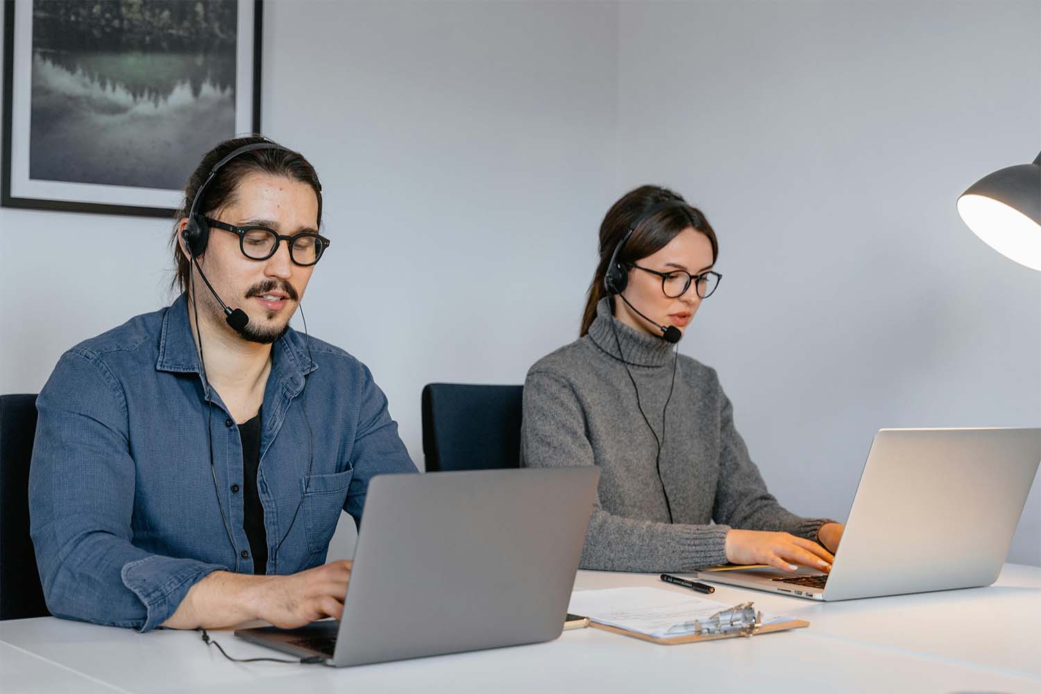 Two people wearing headsets working on laptops at a desk in an indoor setting.