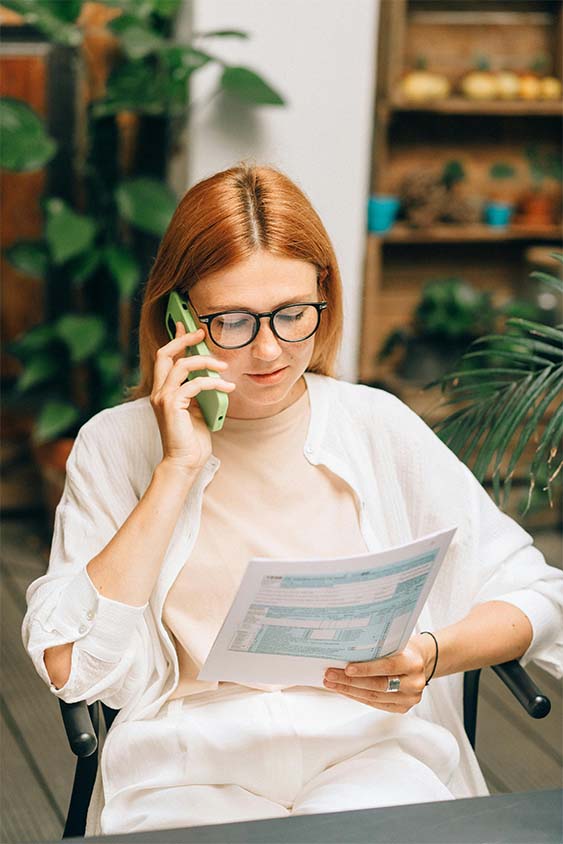 Woman with red hair and glasses talking on a green phone while holding a document.
