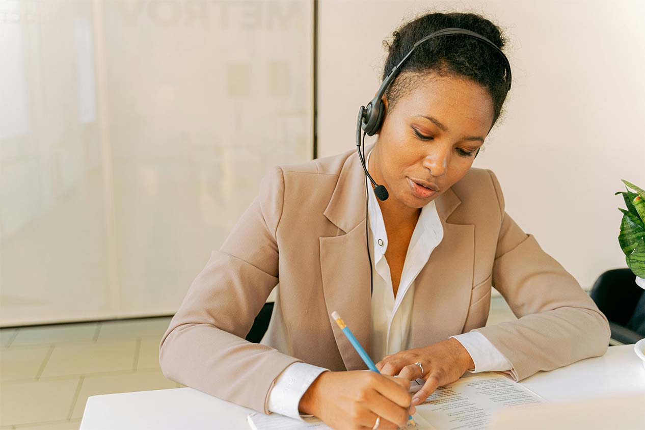A woman with a headset writing notes on a notepad at a desk.